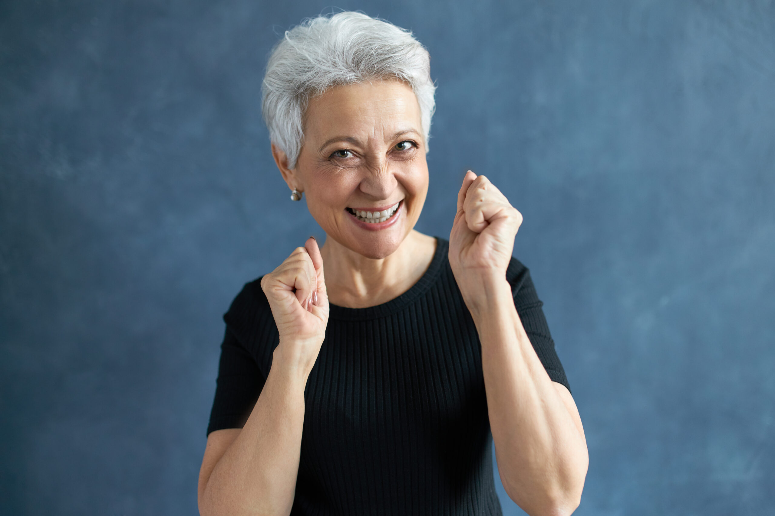 Portrait of overjoyed happy female pensioner with stylish haircut looking at camera with cheerful broad smile, clenching fists, expressing excitement. Human facial expressions and reaction